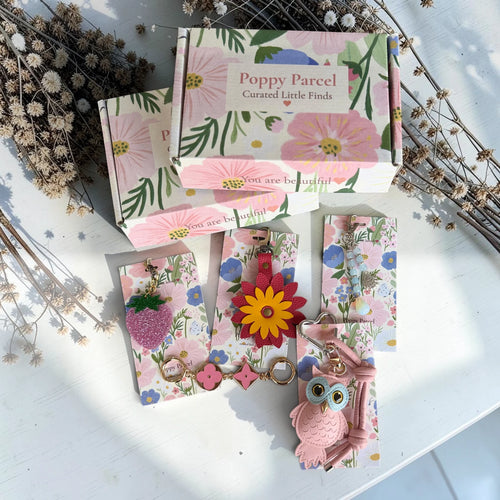 Poppy Parcel classic bag charm gift set styled on a rustic white backdrop with floral boxes, strawberry charm, leather flower charm, owl keychain, and beaded charm.