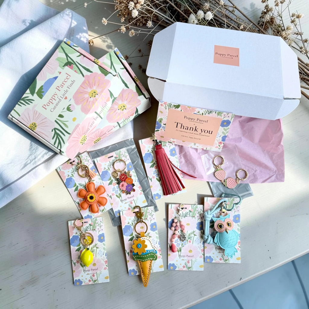 Curated bag charm gift set from Poppy Parcel displayed beside its floral mystery box packaging, featuring colorful keychains, a tasseled charm, and a thank-you card