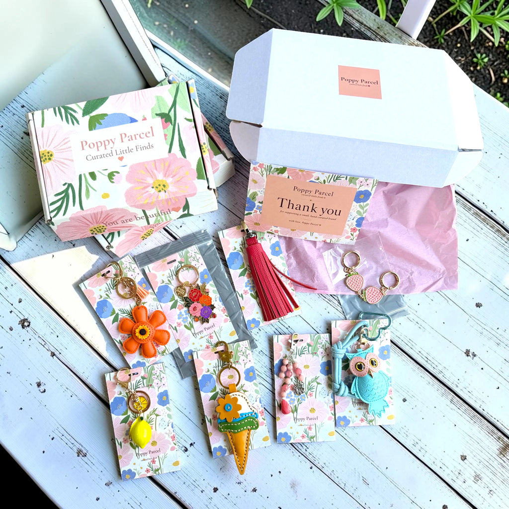 Open Poppy Parcel Deluxe mystery box with floral packaging, thank-you card, and assorted bag charms including a flower, owl, ice cream cone, and beaded keychain laid out on a wooden surface